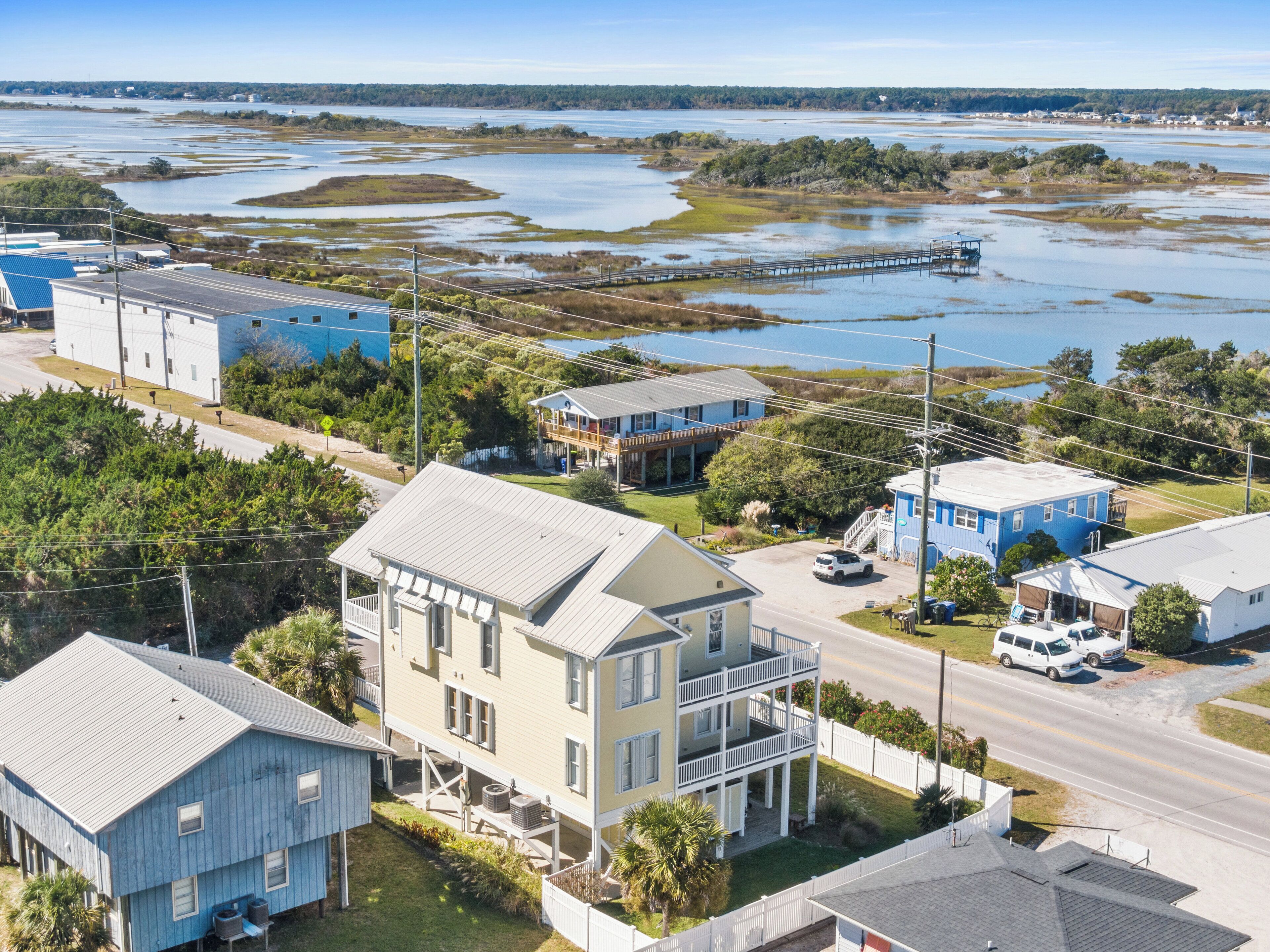 A coastal town with a prominent white house with a balcony, a blue building, and a dock extending into a bay.