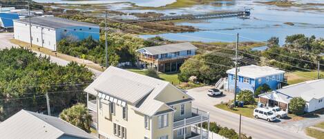 A coastal town with a prominent white house with a balcony, a blue building, and a dock extending into a bay.