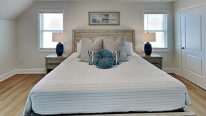 A bedroom with a large bed, two nightstands, a wooden headboard, two windows with blinds, a door, and a framed picture on the wall. - Pelican Landing by Topsail Realty (Sneads Ferry)