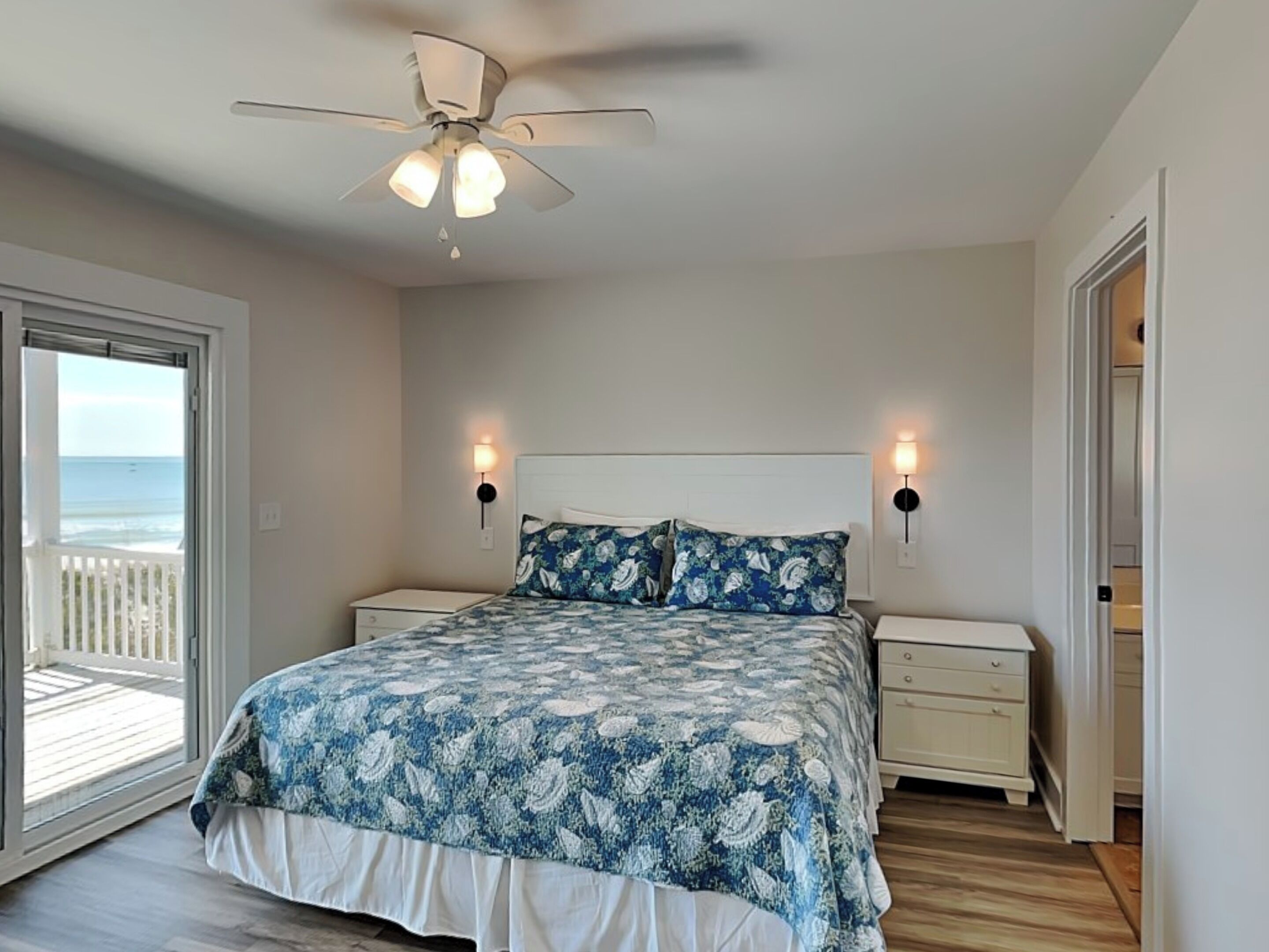 A bedroom with a bed, two bedside tables, a ceiling fan, and a view of the ocean through a sliding door.