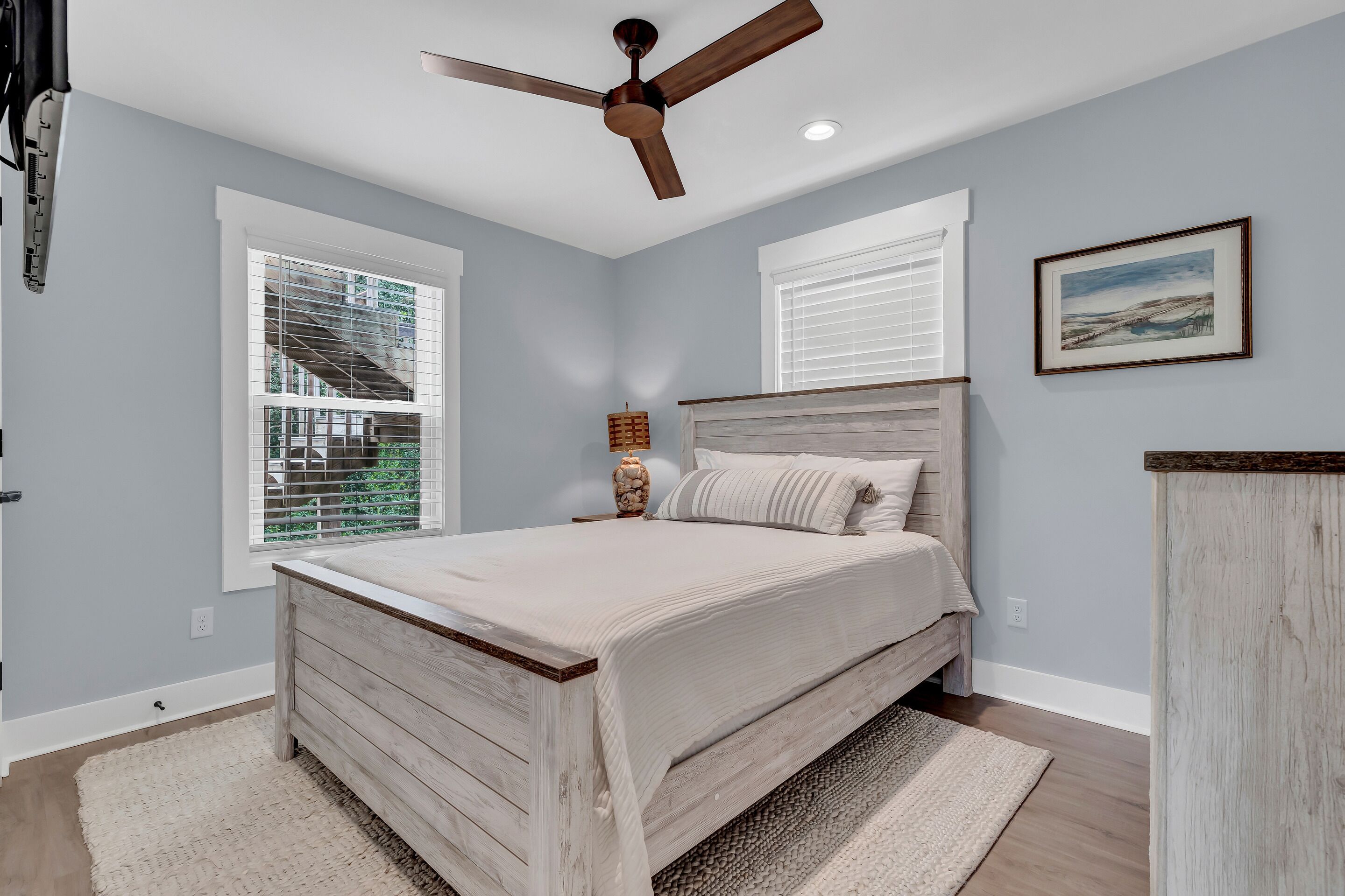 A bedroom with a bed, a ceiling fan, two windows with blinds, a bedside lamp, and a framed picture on the wall.