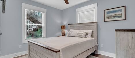 A bedroom with a bed, a ceiling fan, two windows with blinds, a bedside lamp, and a framed picture on the wall.
