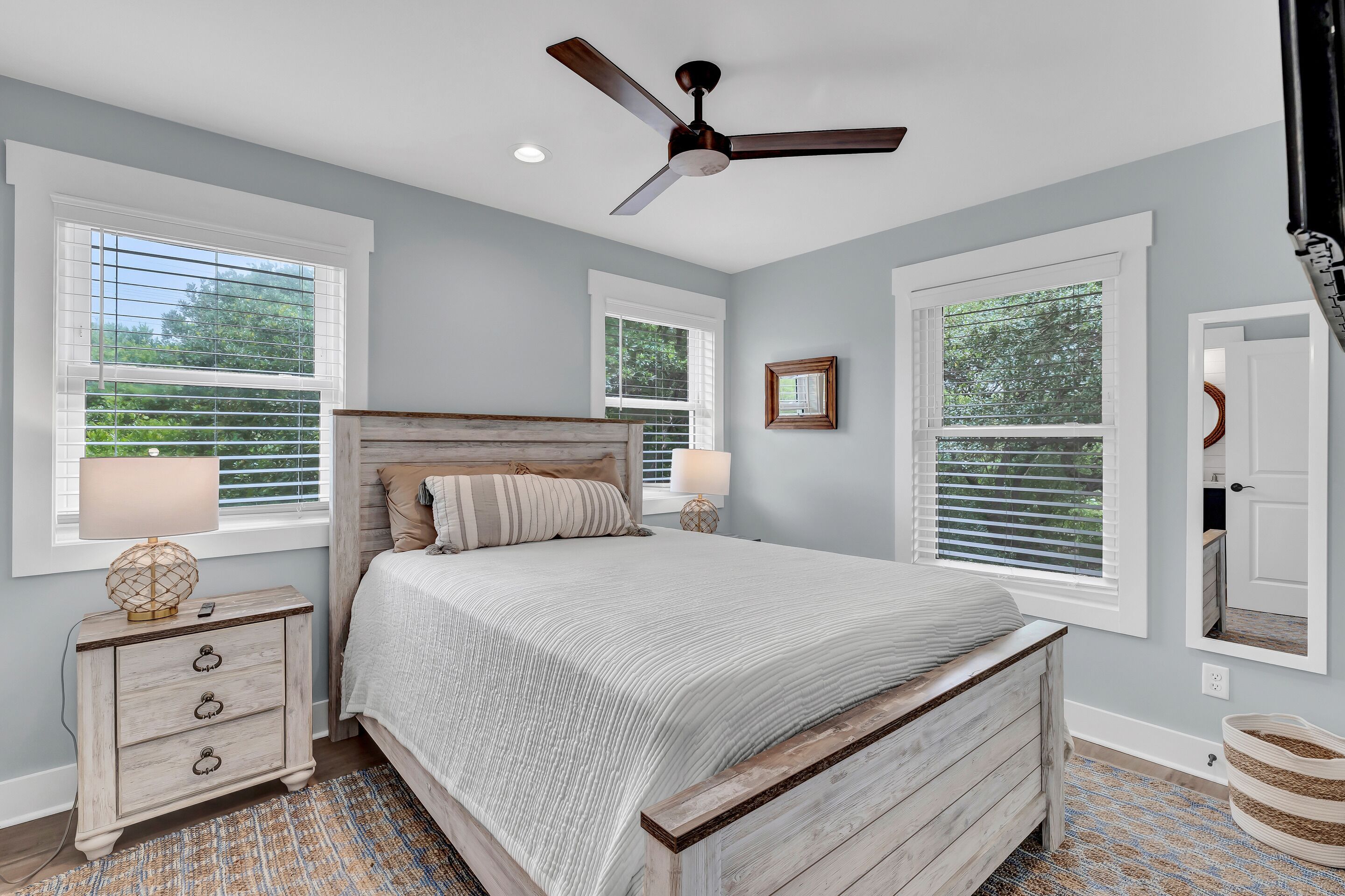 A bedroom with a wooden bed, two bedside tables, a ceiling fan, and large windows with blinds.