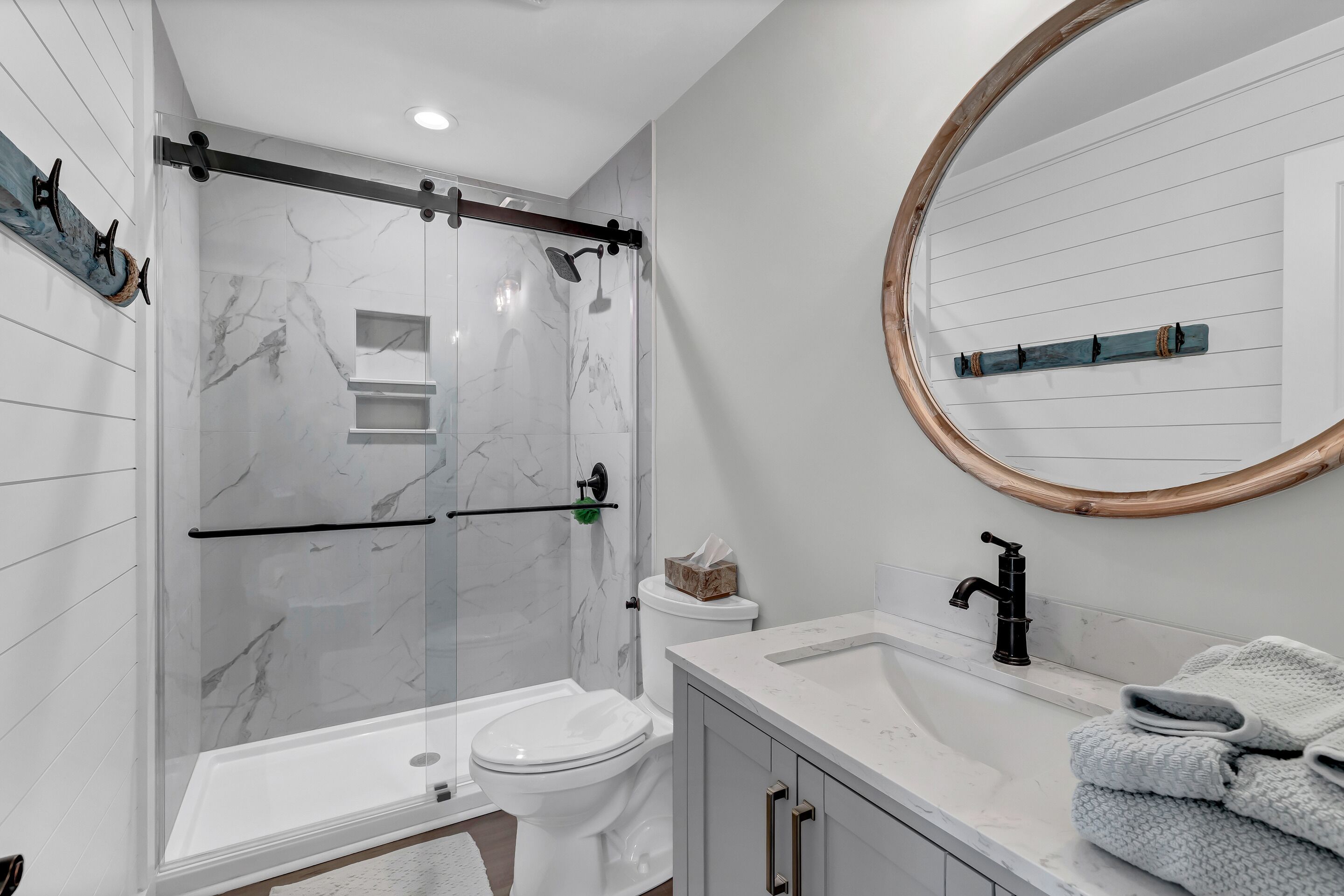 A modern bathroom with a glass-enclosed shower, a white toilet, a sink with a wooden countertop, and a round mirror with a wooden frame.