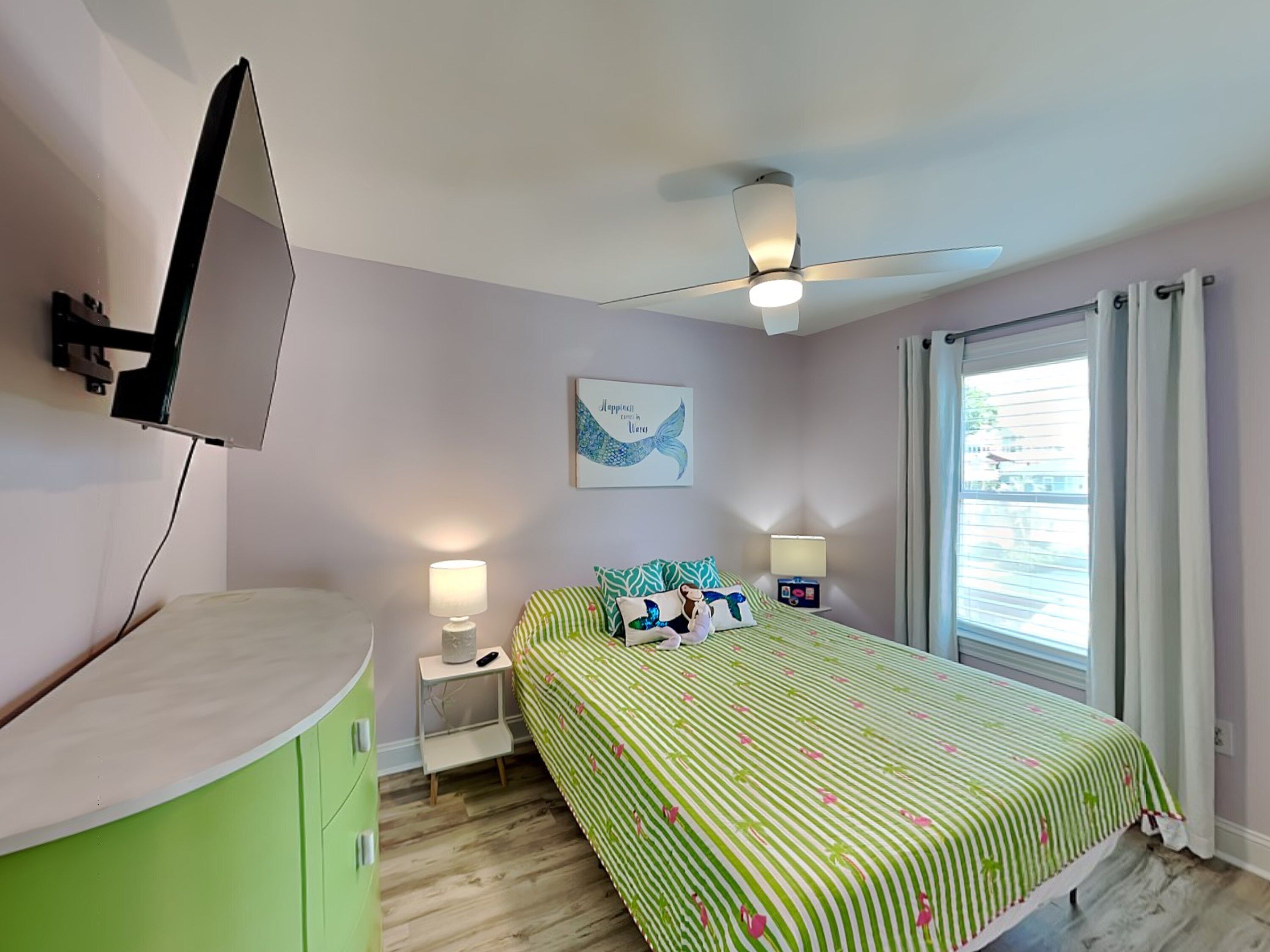 Un dormitorio con cama, escritorio, ventilador de techo, ventana con cortinas y televisor montado en la pared.