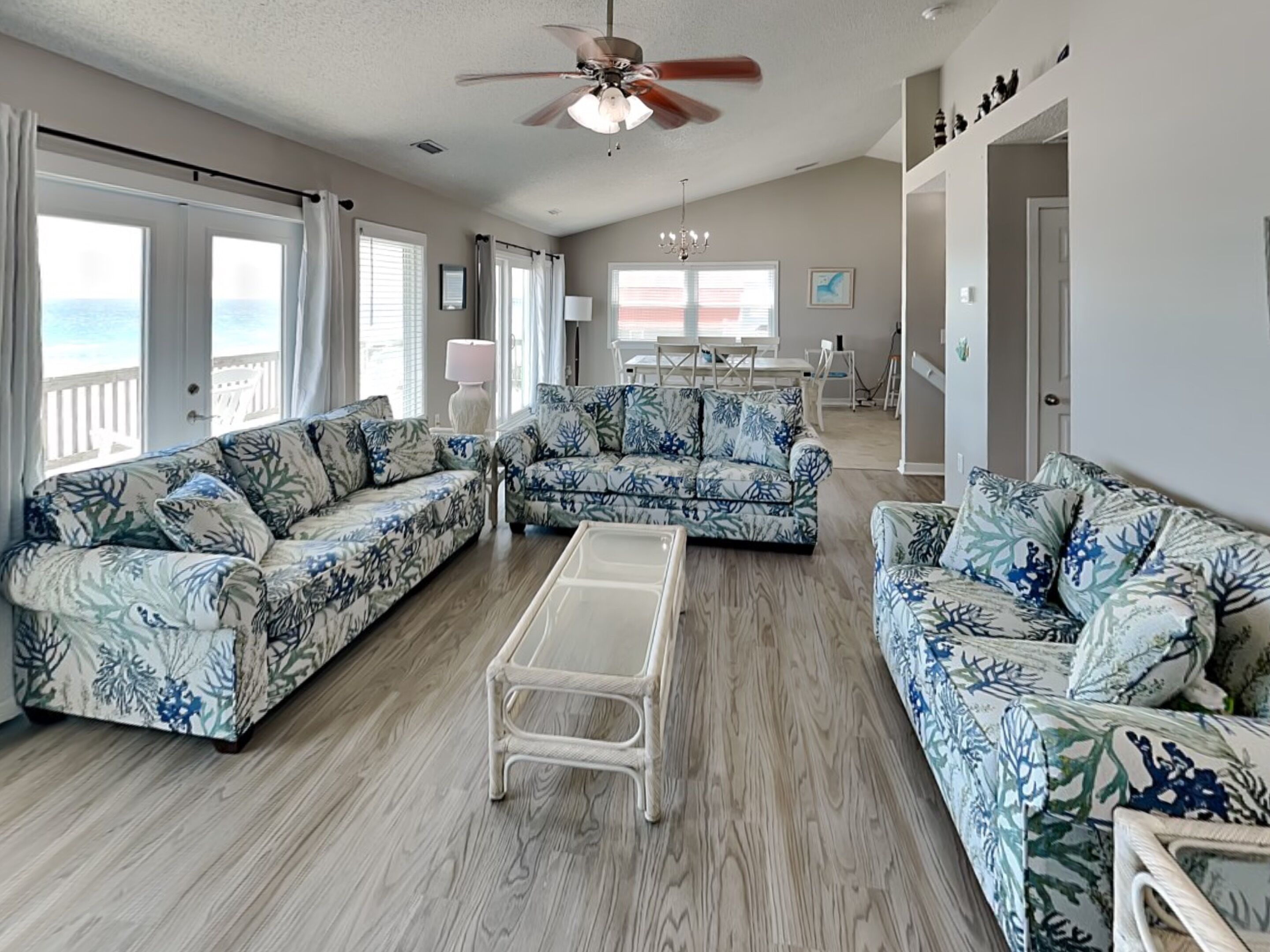 A spacious living room with a ceiling fan, two sofas with a tropical print, a coffee table, and a view of the ocean through large windows.