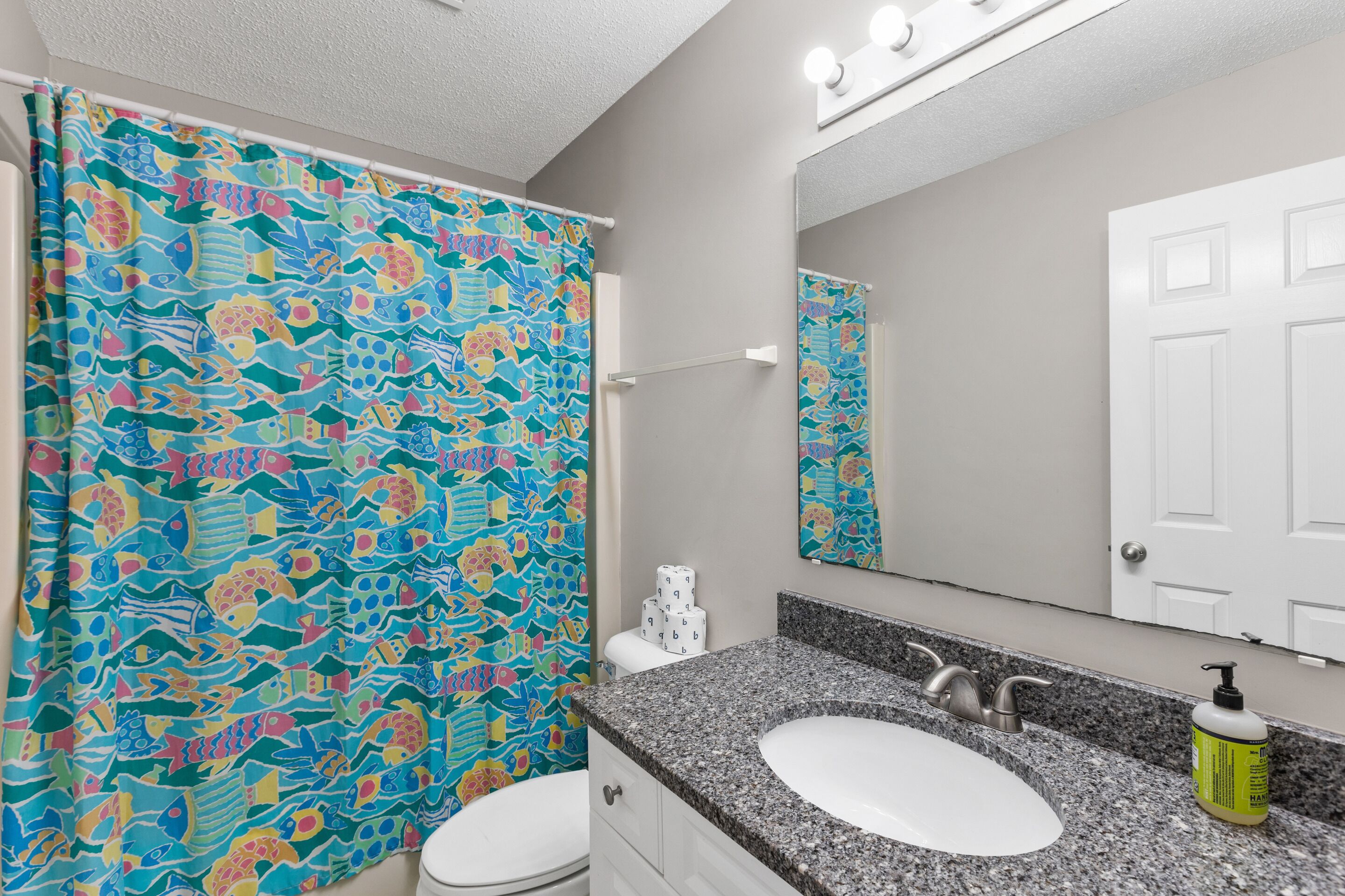 A bathroom with a colorful shower curtain, a white toilet, a granite countertop sink, a mirror, and a white door.