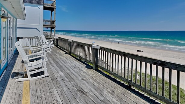 A wooden deck with white chairs, overlooking a beach with clear blue water. - Renegade by Topsail Realty (Holly Ridge)