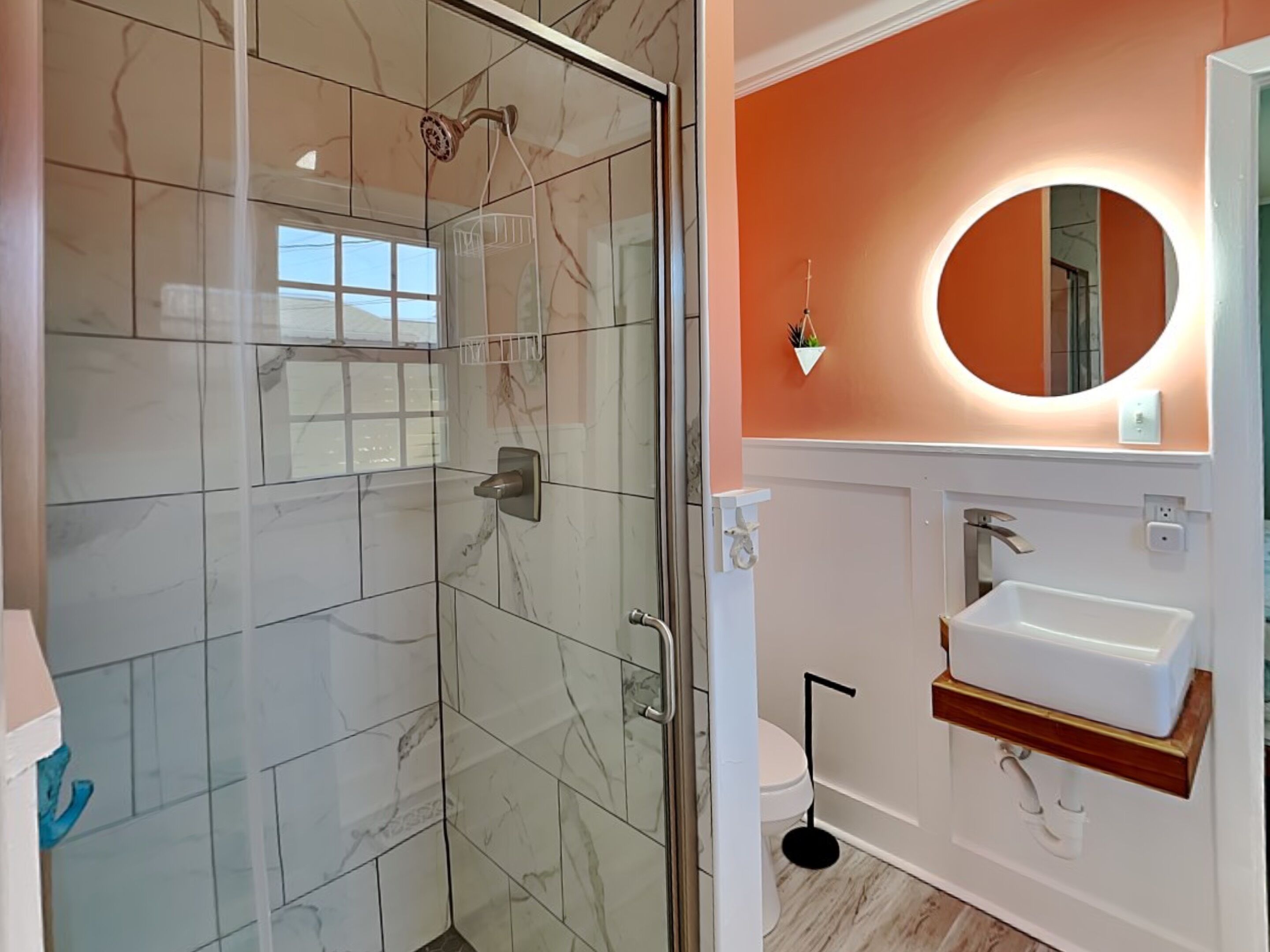 Un baño con ducha envuelta en vidrio, un lavamanos blanco con repisa de madera y un espejo redondo con luz.