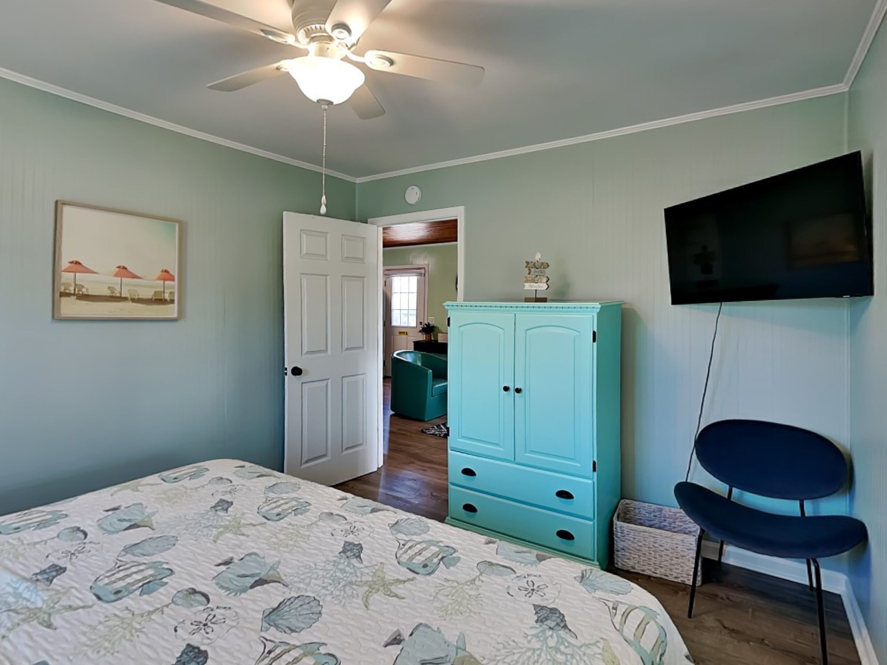A bedroom with a bed, a ceiling fan, a teal cabinet, a TV, and a chair.