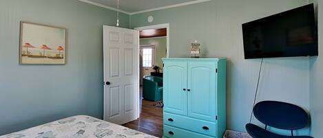 Une chambre à coucher avec un lit, un ventilateur de plafond, une commode de couleur turquoise, un téléviseur et une chaise.