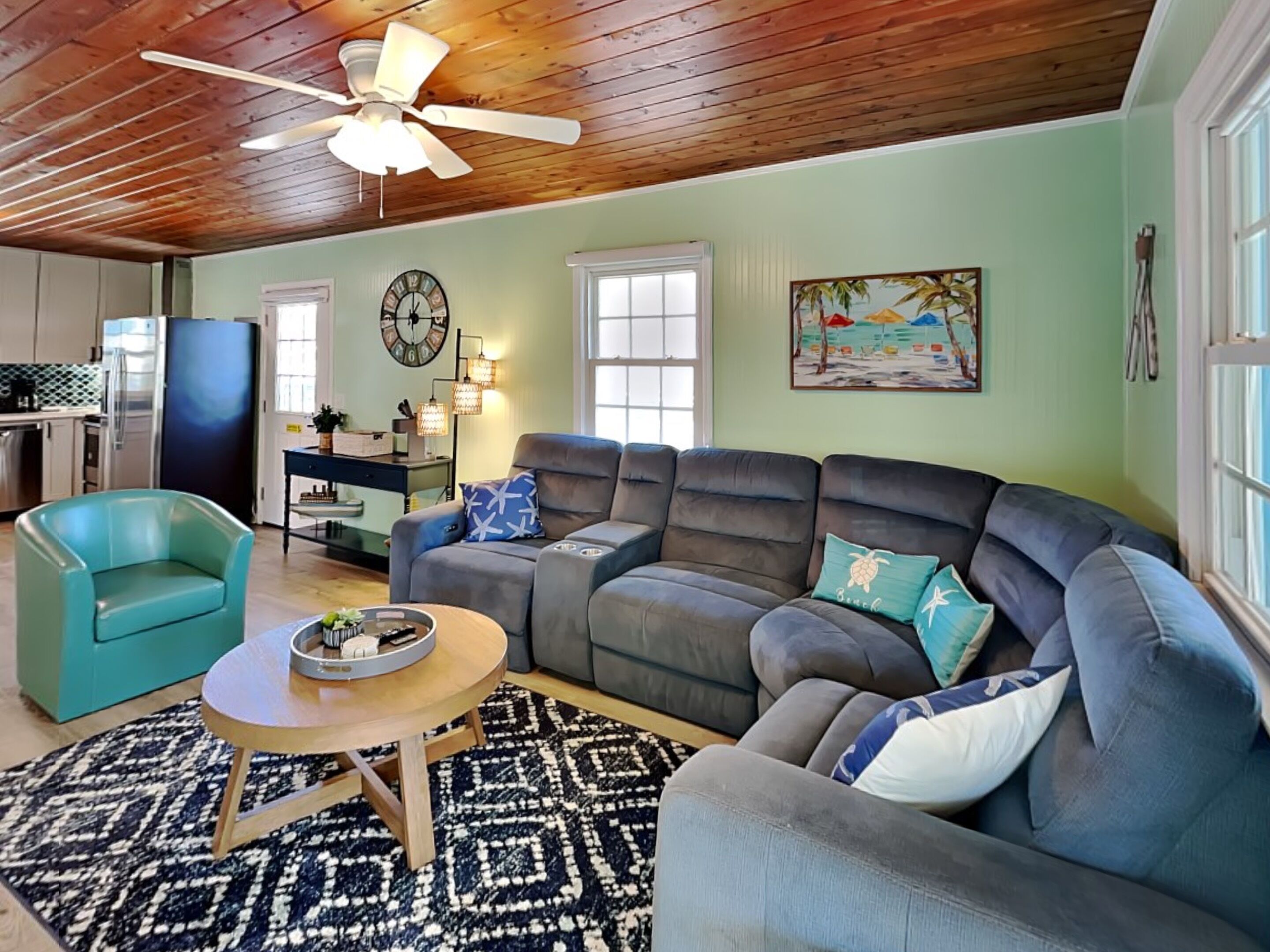 A living room with a sofa, a teal armchair, a wooden coffee table, a patterned rug, a ceiling fan, a clock, a painting, and a kitchen area with a refrigerator and a stove.