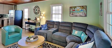 A living room with a sofa, a teal armchair, a wooden coffee table, a patterned rug, a ceiling fan, a clock, a painting, and a kitchen area with a refrigerator and a stove.