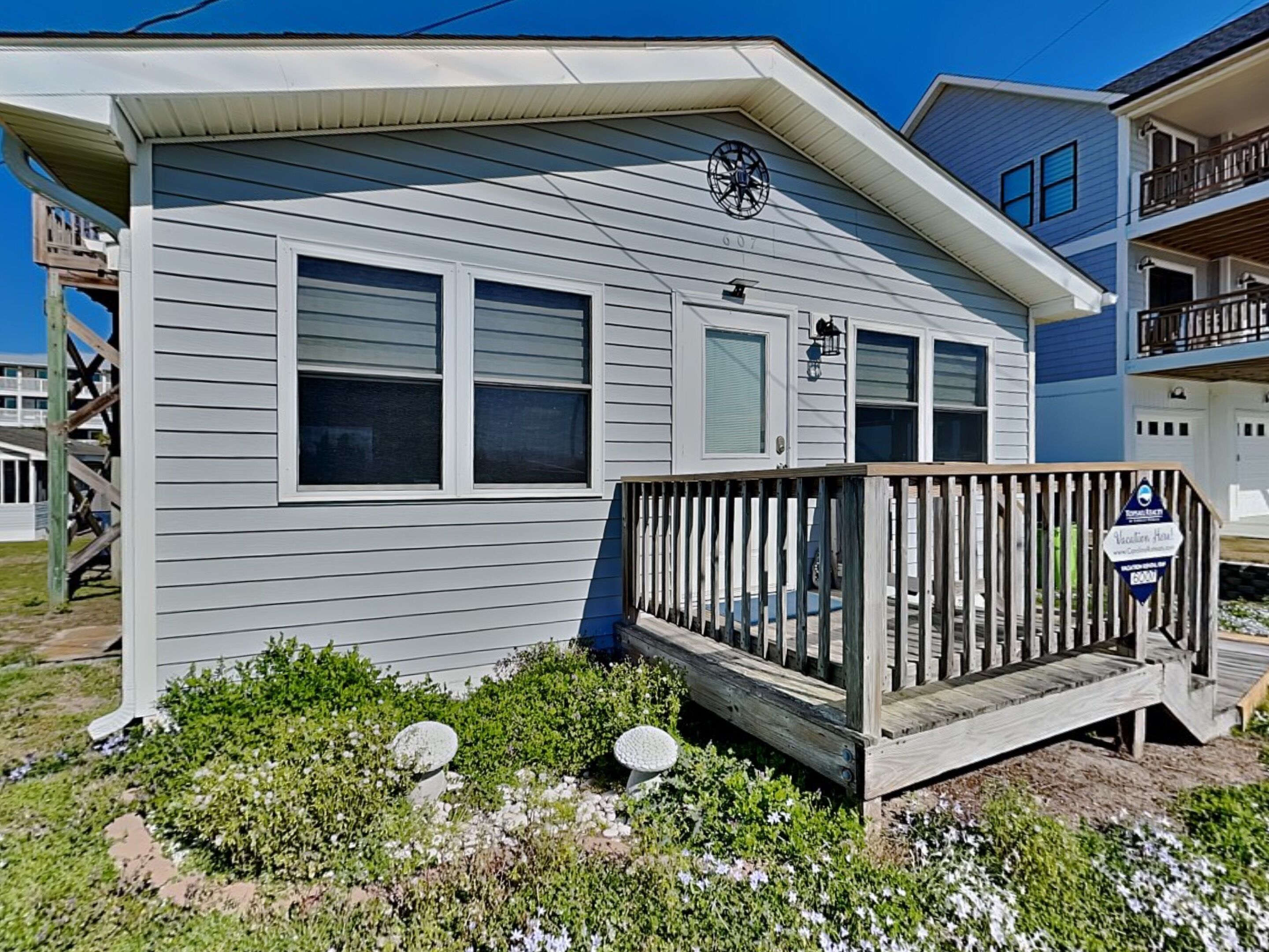 A two-story house with a front deck, a mailbox, and a small garden with flowers and shrubs.