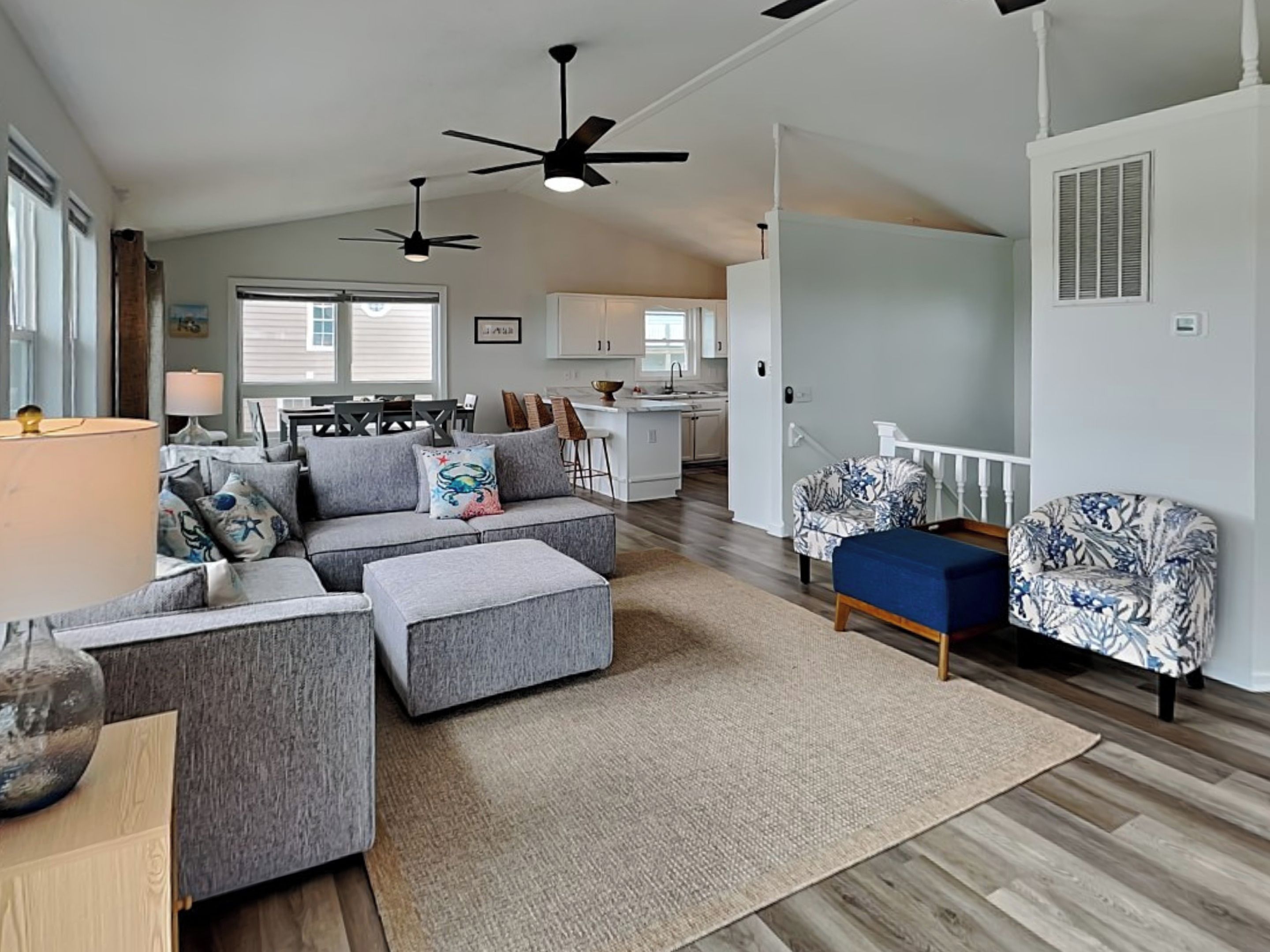Un espacio habitable amplio con un sofá seccional gris, un taburete azul y sillones estampados. Se ve una cocina al fondo y hay ventiladores de techo.