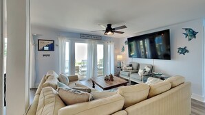 Un salon avec un canapé beige, une table basse, un téléviseur fixé au mur et un ventilateur de plafond.