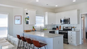 A modern kitchen with white cabinetry, a central island with bar stools, stainless steel appliances, and a breakfast bar. - Sandy By The Sea by Topsail Realty (Sneads Ferry)