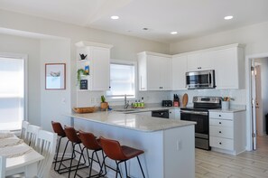 A modern kitchen with white cabinetry, a central island with bar stools, stainless steel appliances, and a breakfast bar. - Sandy By The Sea by Topsail Realty (Sneads Ferry)