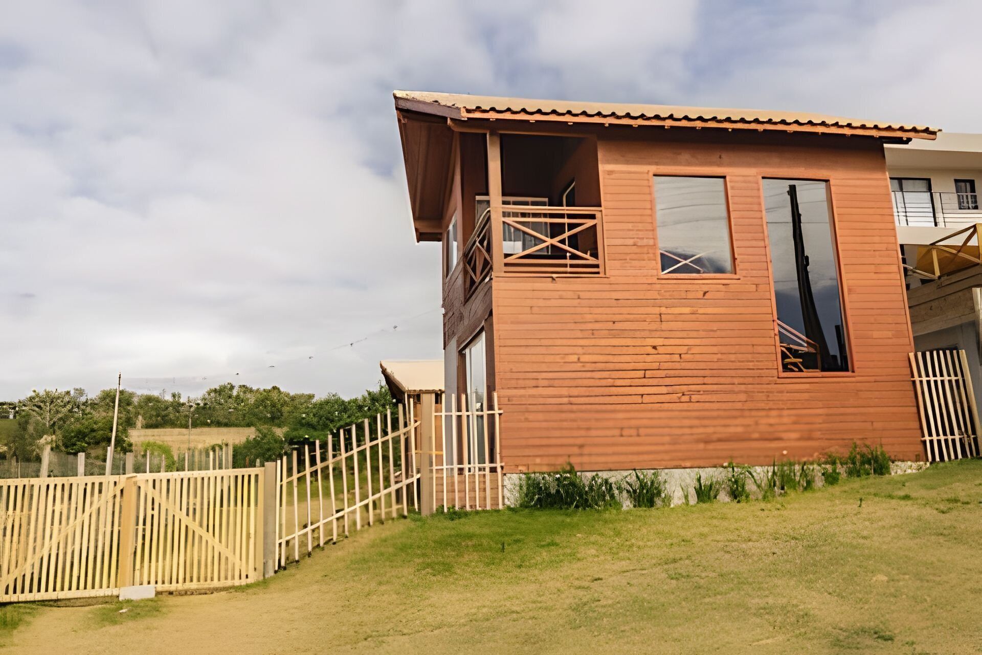A wooden house with a balcony, a fenced yard, and a cloudy sky.