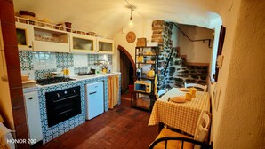 Una cocina compacta con estufa, horno y refrigerador, una mesa de comedor con mantel a cuadros y una escalera de piedra que lleva a un nivel superior.