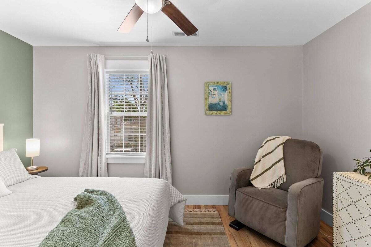 Quarto com cama, cadeira, escrivaninha, janela com cortinas, ventilador de teto e um quadro na parede.