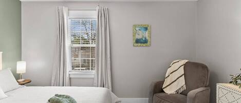 Quarto com cama, cadeira, escrivaninha, janela com cortinas, ventilador de teto e um quadro na parede.