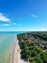 An aerial view of a coastal area with a beach, dense greenery, and a clear blue sky.