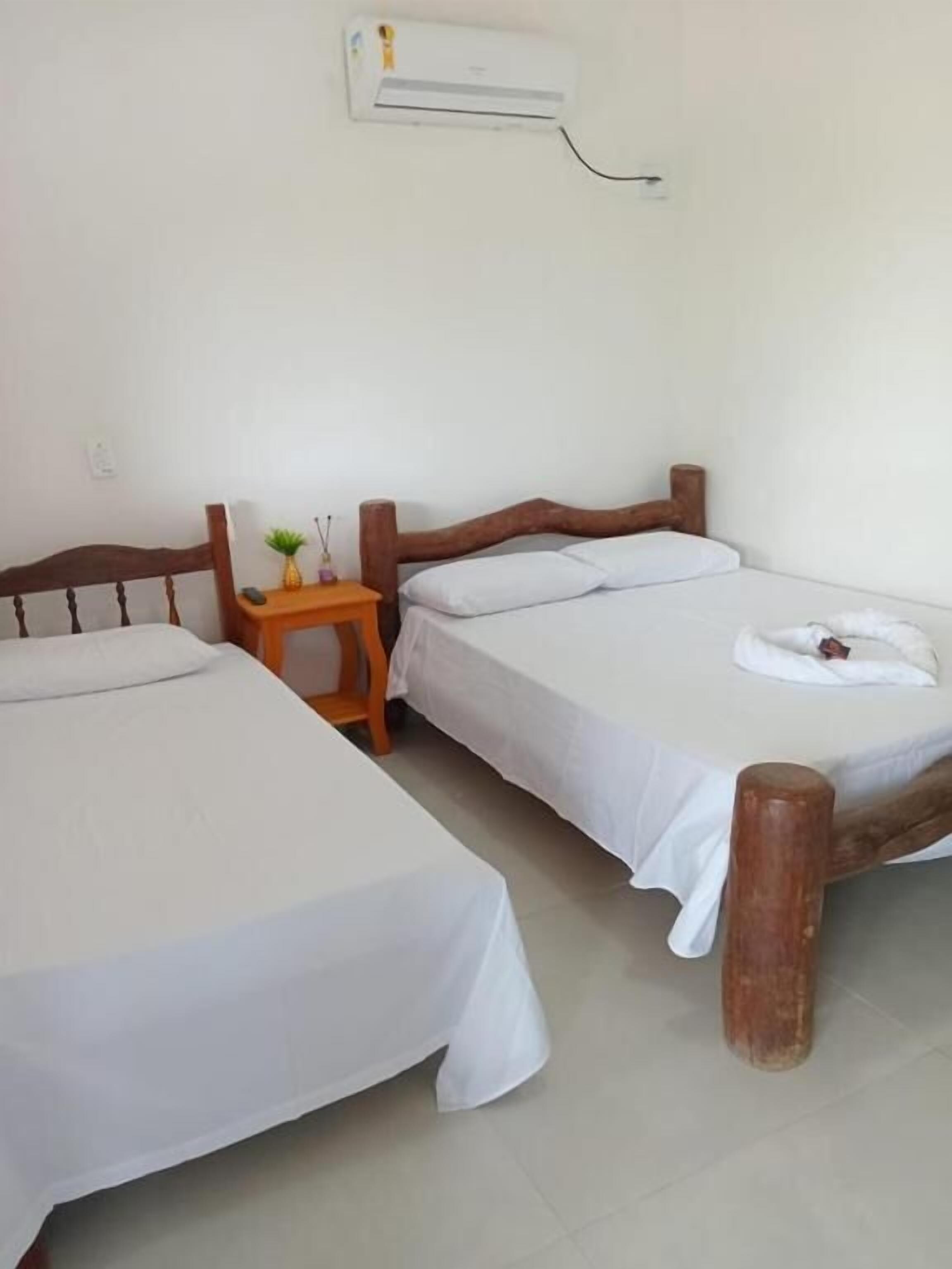 A room with two single beds, a wooden nightstand with a small plant, and a wall-mounted air conditioner.