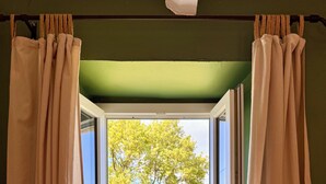 Une fenêtre avec des rideaux de couleur beige, donnant sur un arbre et de l’herbe.