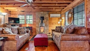 Un salon chaleureux, aménagé dans le style d’un chalet, avec deux canapés en cuir, un pouf rouge, une cheminée en pierre, un téléviseur et un ventilateur de plafond.
