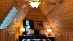 Une chambre confortable située sous les toits, avec un plafond en pente, un lit recouvert d’une couverture à carreaux, deux lampes de chevet, un ventilateur de plafond et une fenêtre avec des stores.
