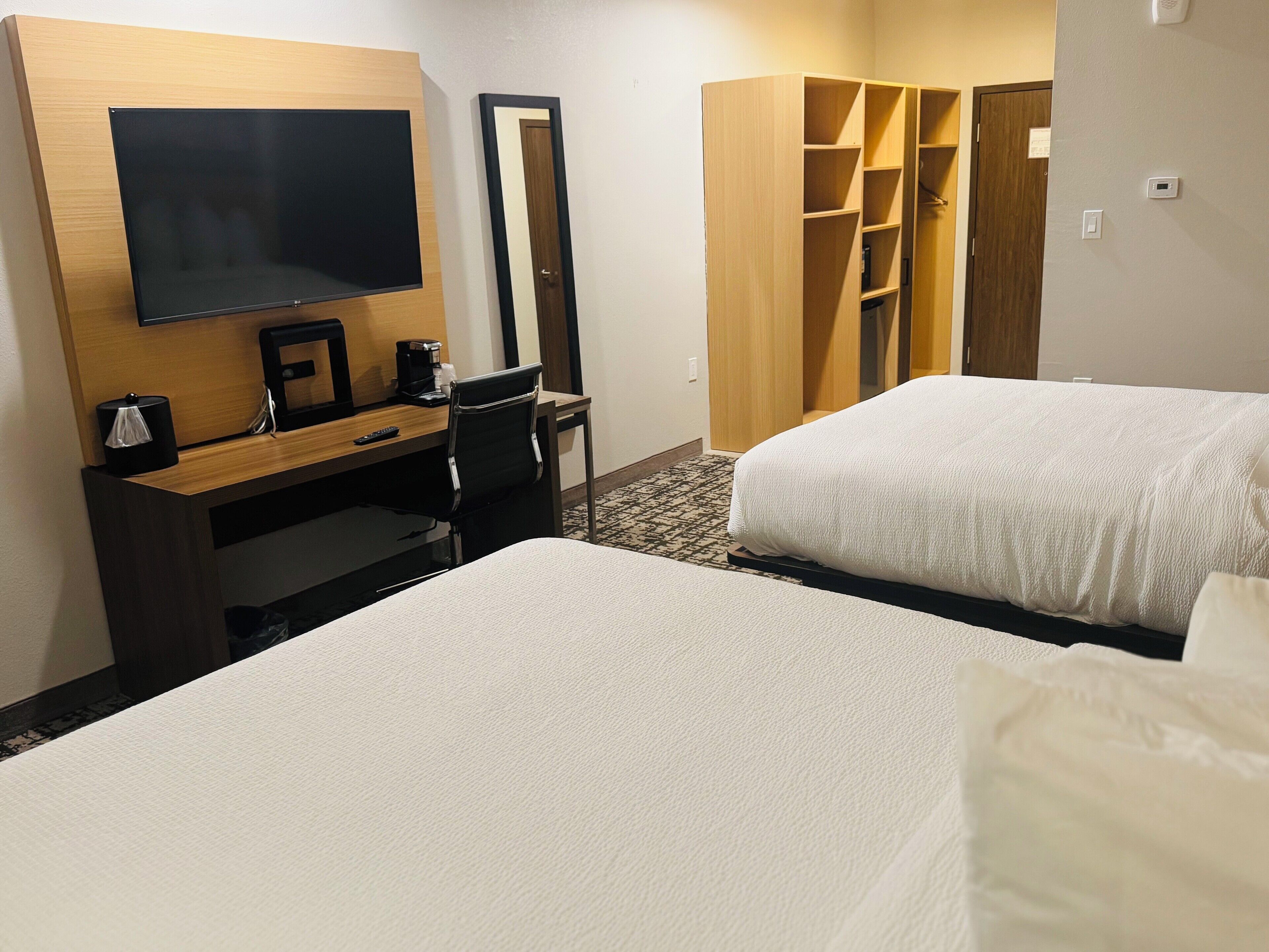 Quarto de hotel com duas camas, uma escrivaninha com cadeira, televisão, guarda-roupa e um armário pequeno.