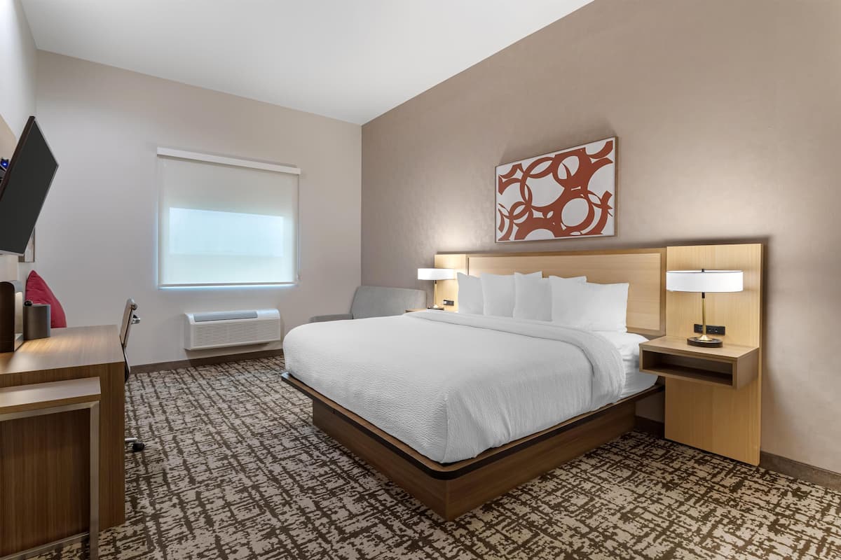 A hotel room with a large bed, bedside lamps, a desk with a flat-screen TV, a window with blinds, and a wall-mounted artwork.