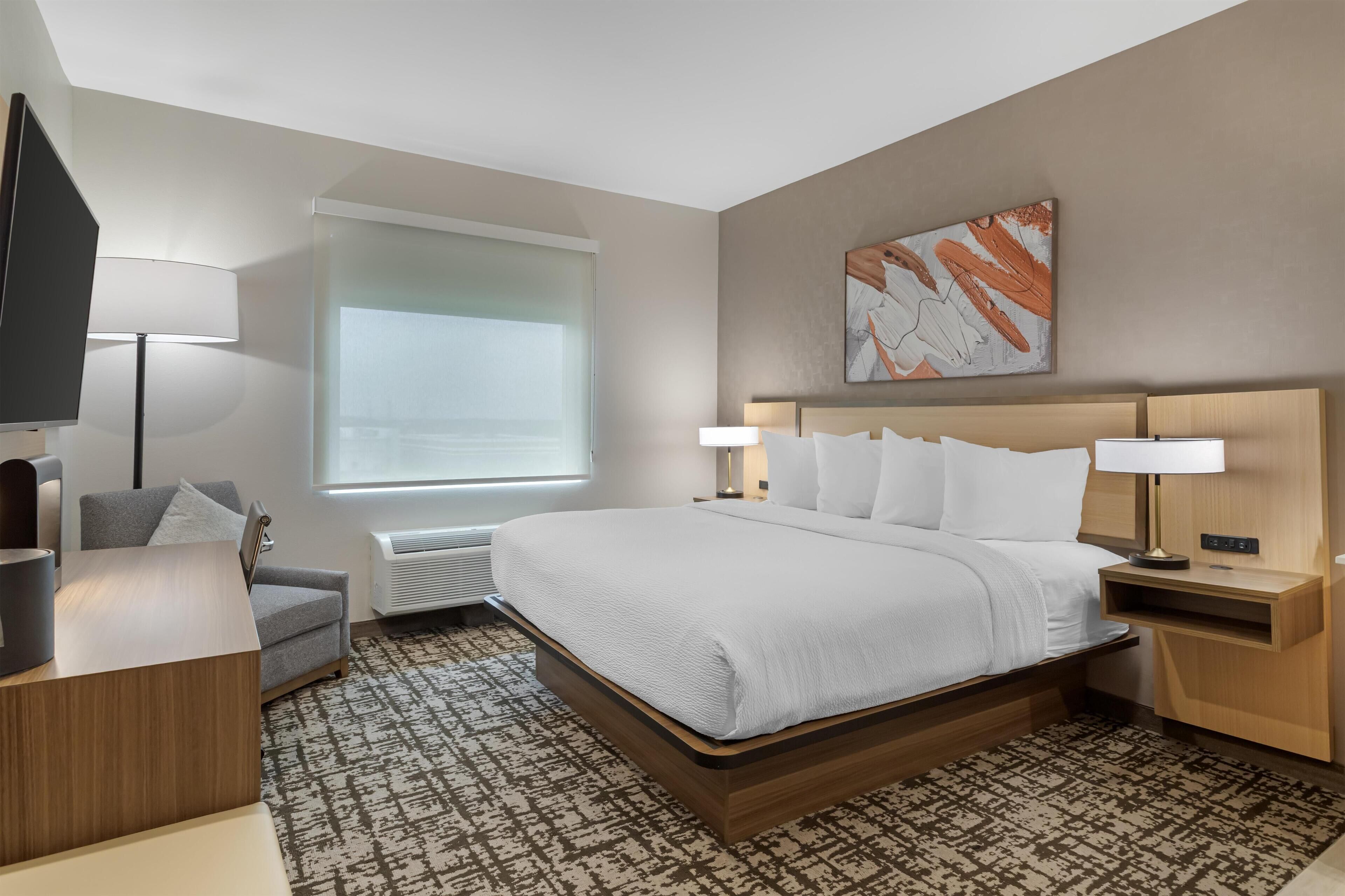 Une chambre d’hôtel moderne avec un grand lit, des tables de chevet avec des lampes, un téléviseur à écran plat, un canapé gris et un tapis à motifs.