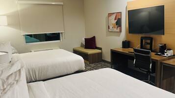 Ein Hotelzimmer mit zwei Betten, einem Schreibtisch mit Stuhl, einem Fernseher und einem Fenster mit Jalousien.