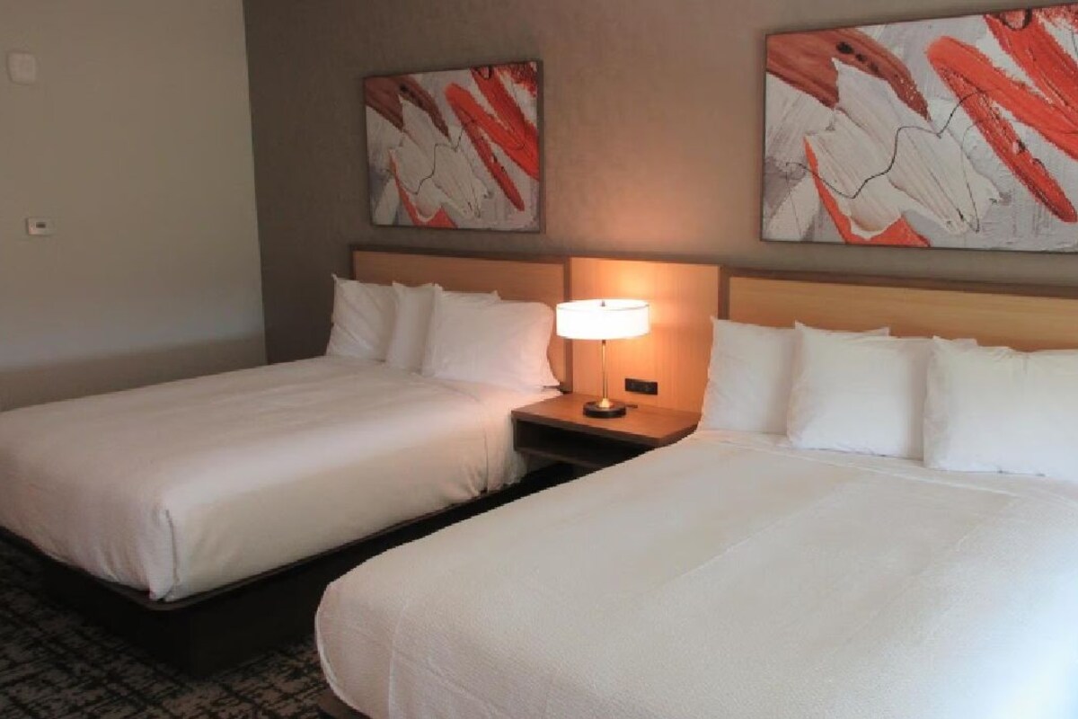 Twee aangrenzende hotelbedden met witte lakens, een nachtkastje met een lamp en abstracte wanddecoratie.