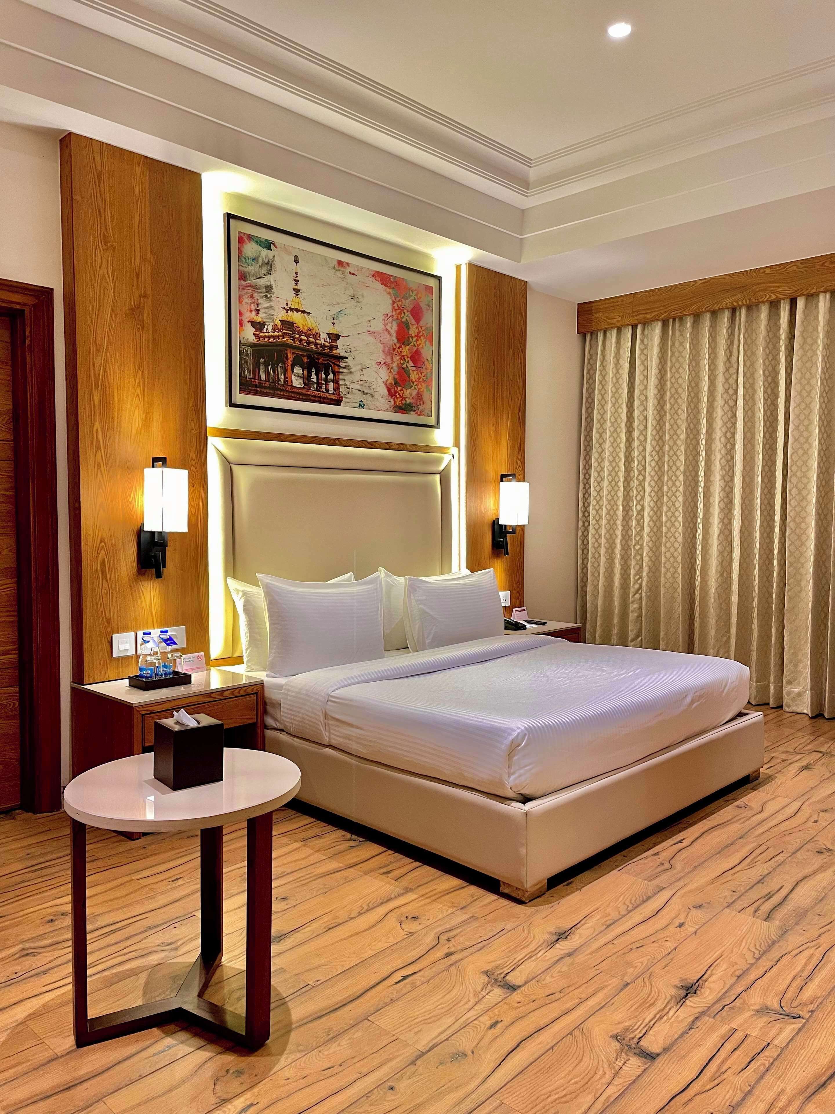 A hotel room with a large bed, a bedside table with a lamp, a small round table, and a wall-mounted painting.