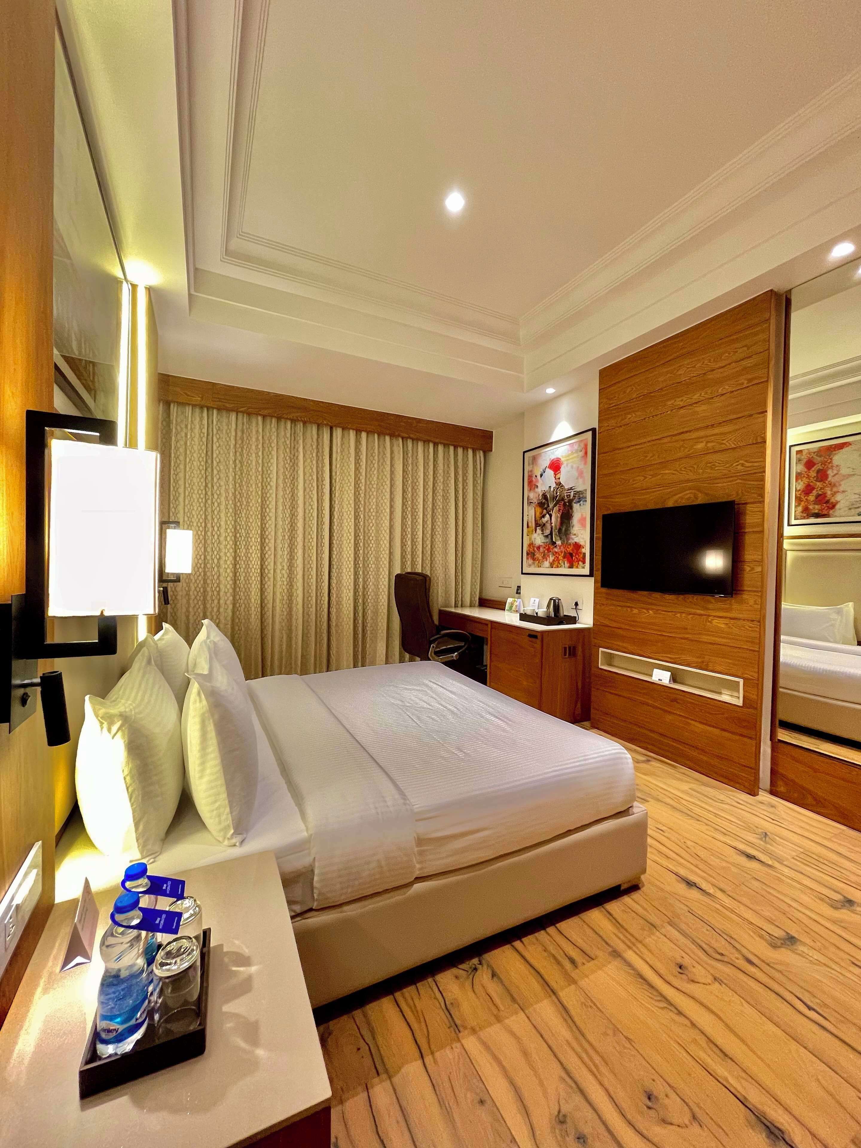 A hotel room with a large bed, a desk with a chair, a flat-screen TV mounted on a wooden panel, and a window with curtains.