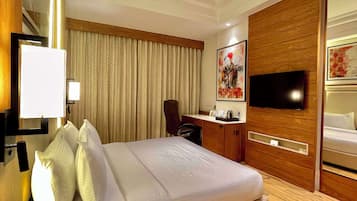 A hotel room with a large bed, a desk with a chair, a flat-screen TV mounted on a wooden panel, and a window with curtains.