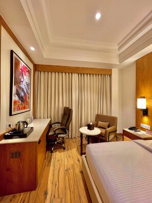 A hotel room with a bed, a desk, a chair, a small table, a lamp, and a painting on the wall.