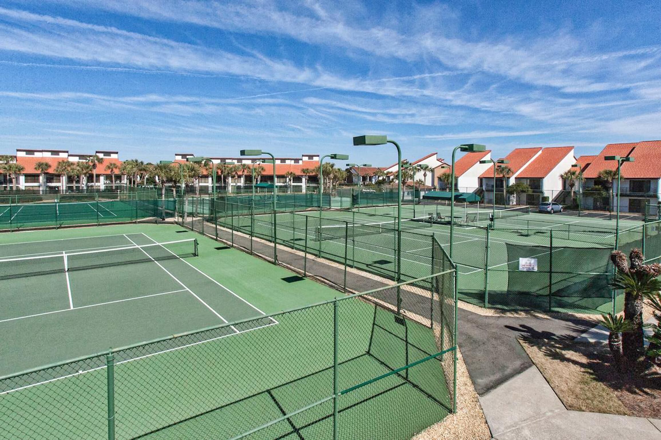 A tennis court complex with multiple courts, surrounded by buildings with red roofs and palm trees.