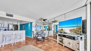 Een moderne keuken met witte kasten, een eethoek met tafel en stoelen, en een grote televisie waarop een strand te zien is.