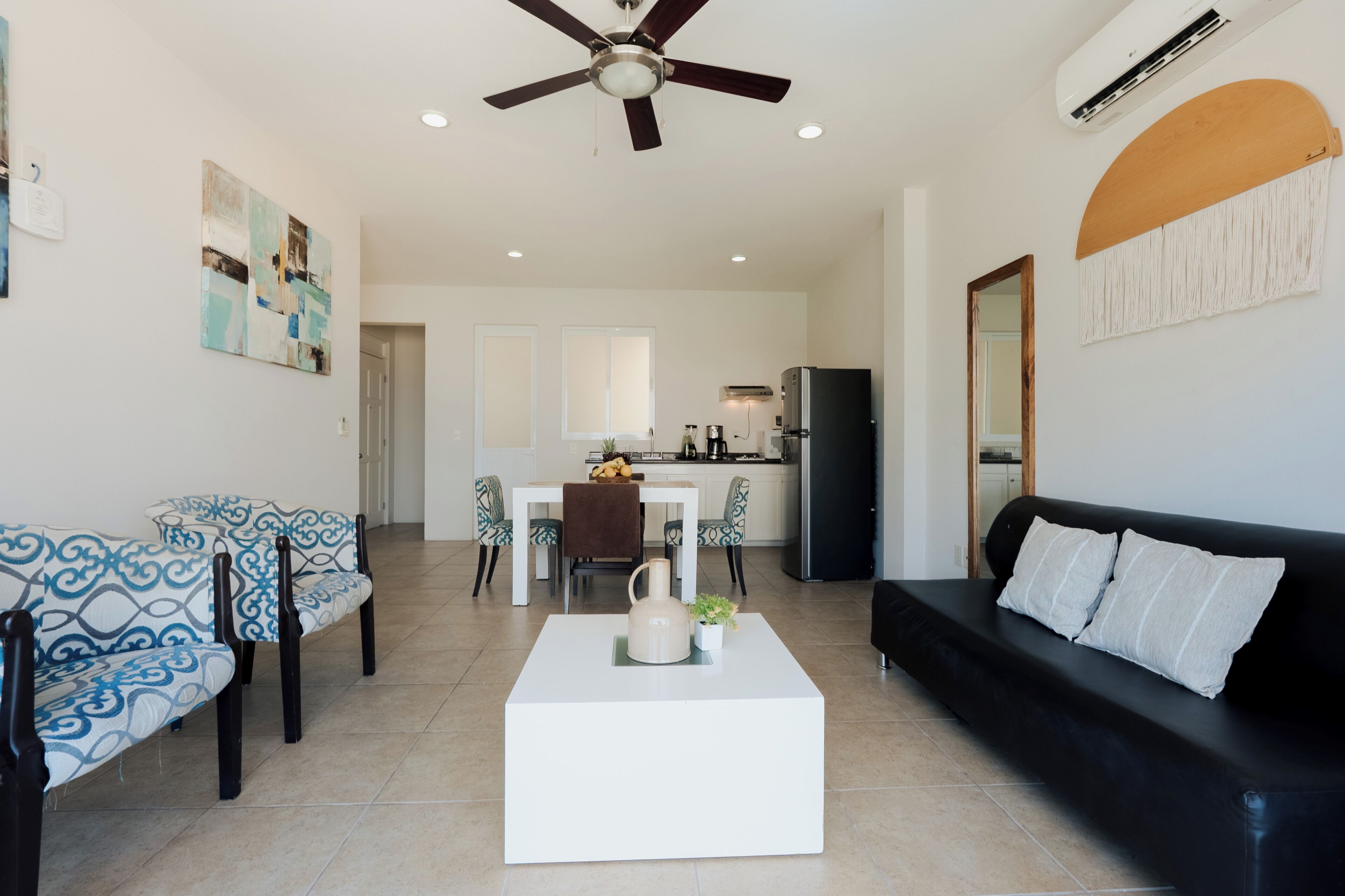 A modern living room with a white coffee table, a black sofa, patterned chairs, a ceiling fan, and a kitchen in the background.