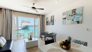 A modern living room with a large window, a dining area with a fruit basket, and a ceiling fan. - Suites La Playa (Santa Maria Huatulco)