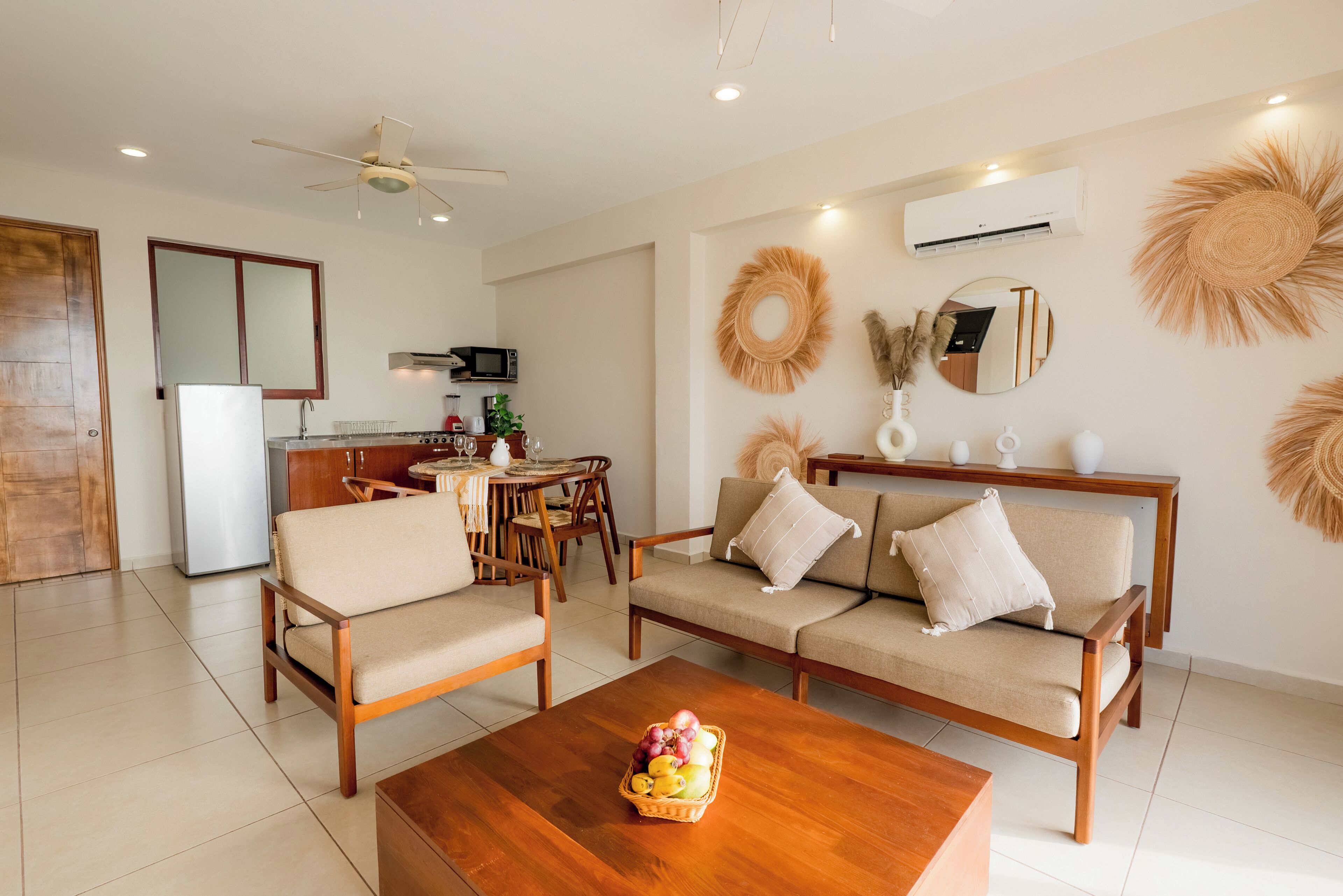 A modern living room with a sofa, chairs, a coffee table with fruit, a dining area with a table and chairs, and a kitchen with appliances.