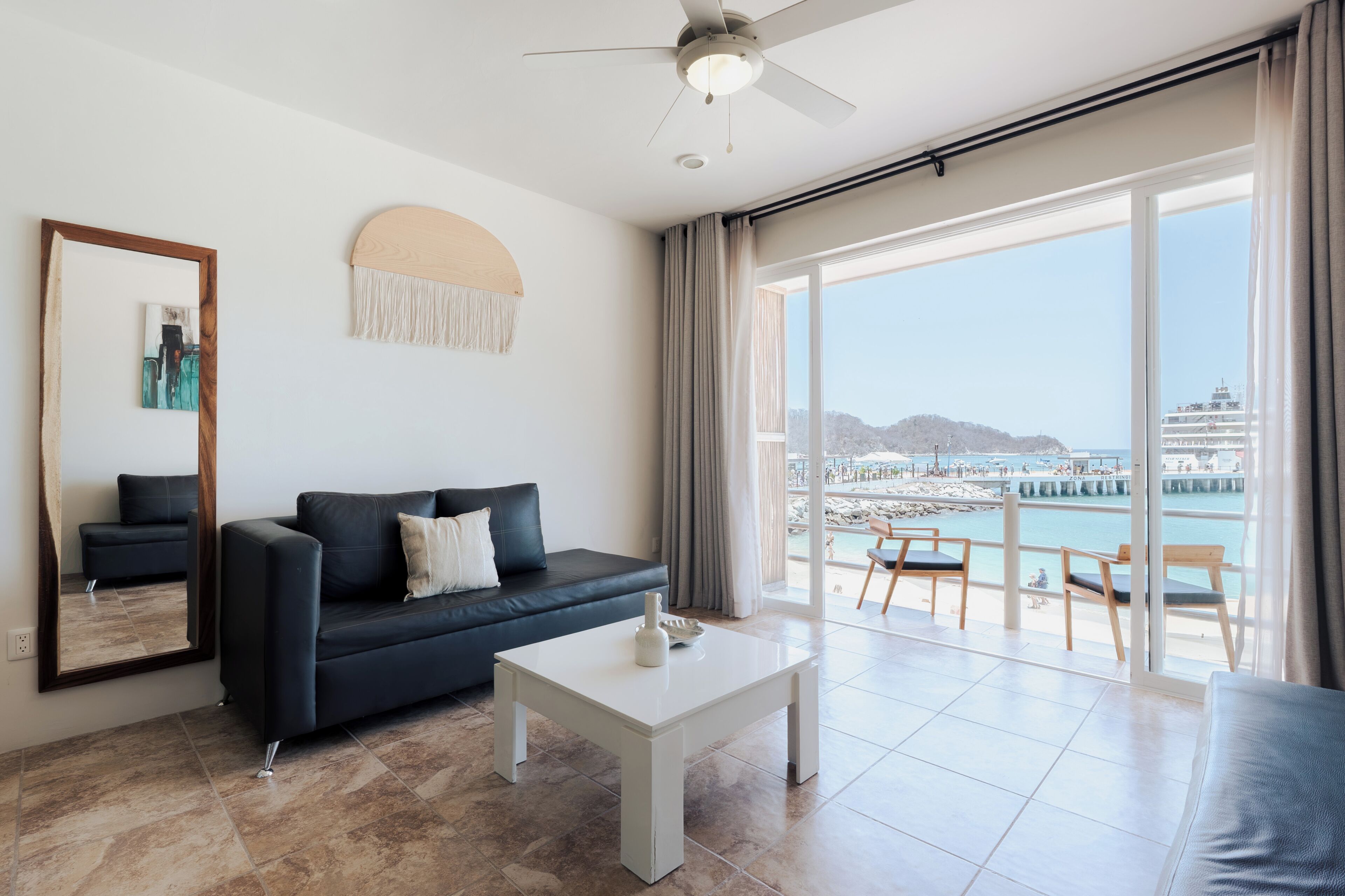 Una sala de estar moderna con un sofá, una mesa de centro, un ventilador de techo, un ventanal con vista a una playa y un muelle, y un espejo en la pared.