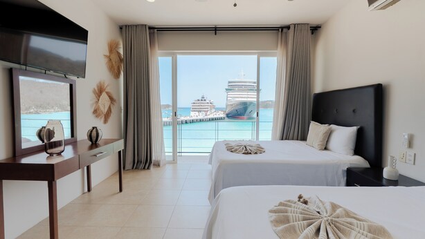 A hotel room with two beds, a TV, a desk, and a view of a cruise ship and pier. - Suites La Playa (Santa Maria Huatulco)