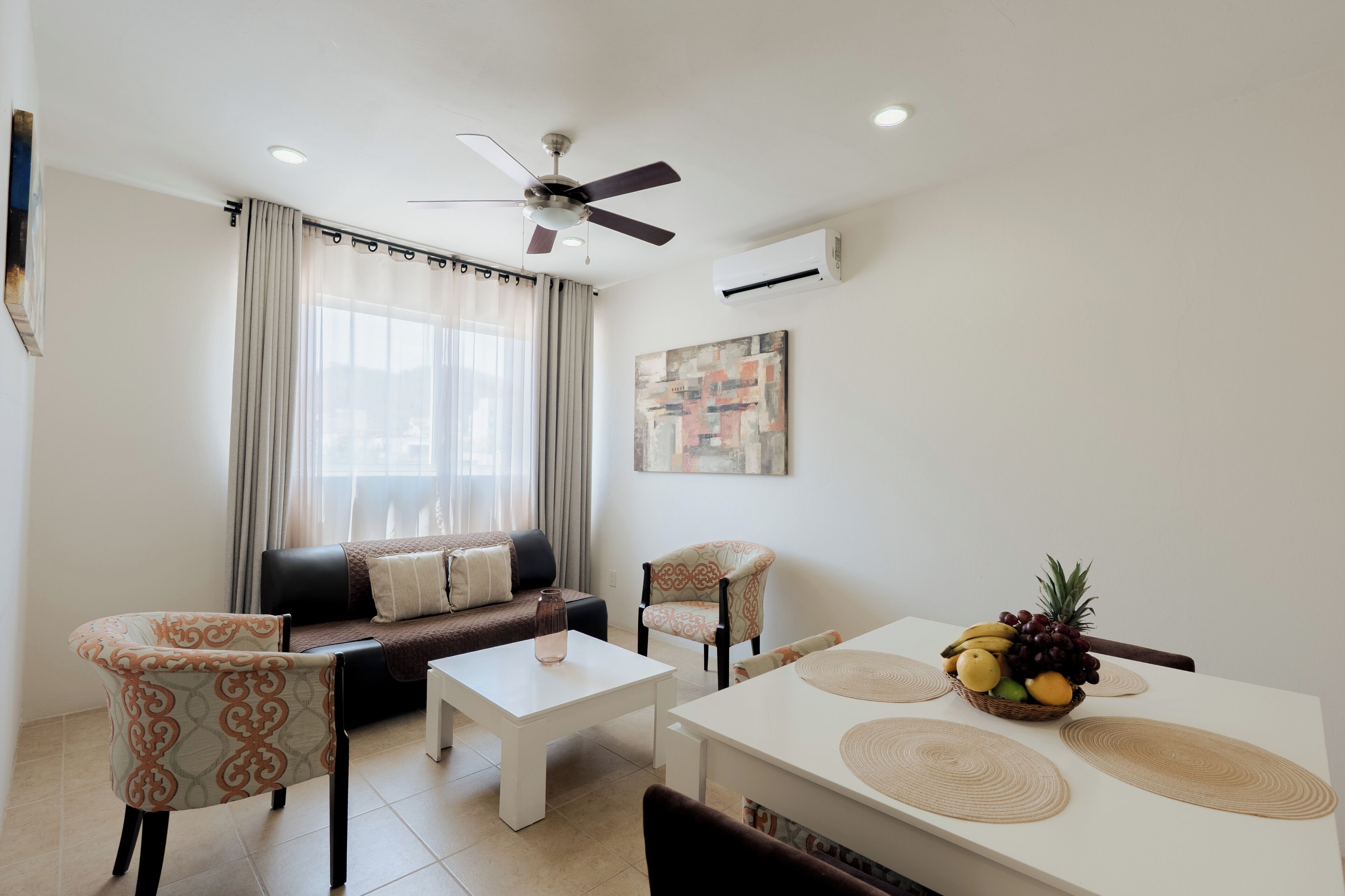A living room with a sofa, two armchairs, a coffee table, a dining table with placemats, a ceiling fan, and an air conditioning unit.