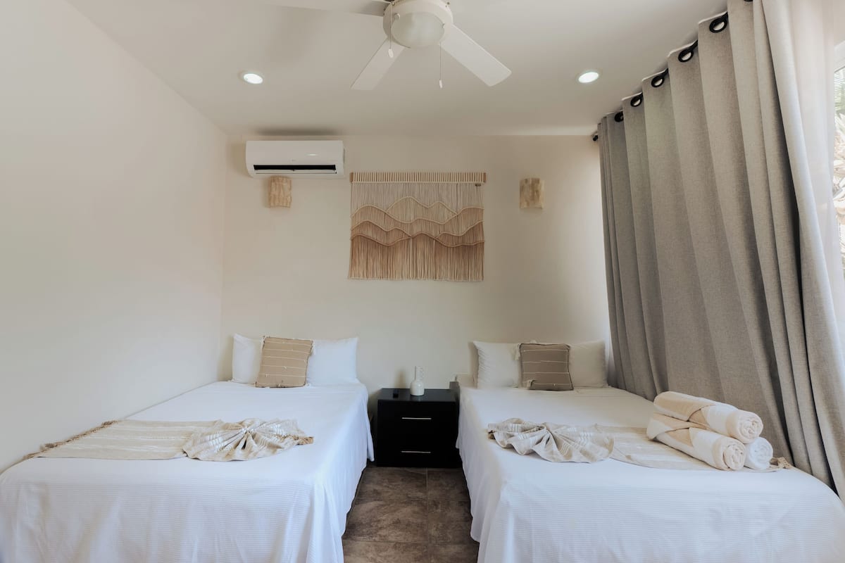 Camera d'albergo con due letti, una ventola a soffitto, un condizionatore d'aria, un pannello decorativo a parete e una finestra con tende.