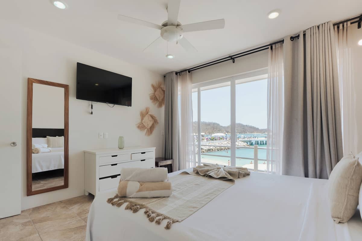 Una camera da letto con un letto, una TV, una ventola a soffitto, una finestra con tende e vista sulla spiaggia.